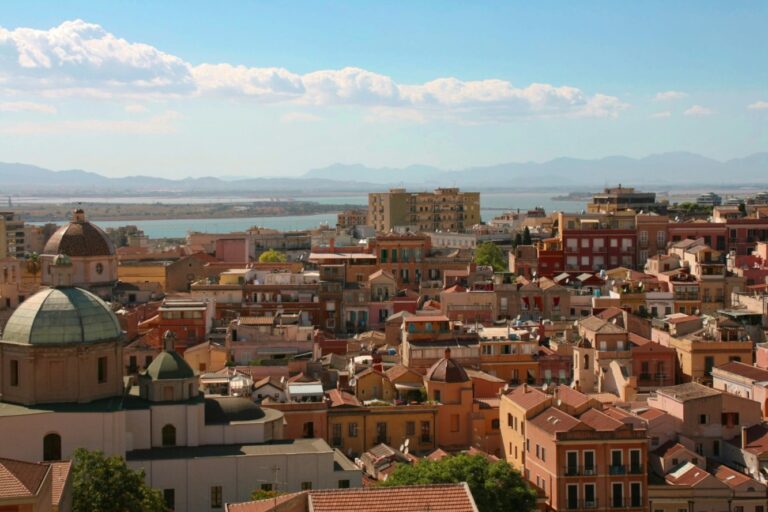 Panoramic view of Cagliari’s historic center with terracotta rooftops, domed churches, and the lagoon stretching toward distant mountains under a clear sky.