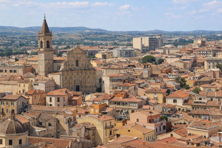 Panoramic view of Sassari with dense terracotta rooftops, the Cathedral of San Nicola rising above the historic center, and rolling hills in the background.