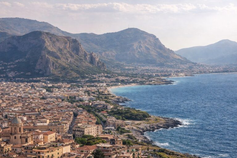 I need you to please generate a high res landscape image for slide 4 of the below, to insert in the "Distinct Territories in Sicilia" section Palermo & Conca d’Oro (Northwest metro gravity) Slide 1 — Why this place exists Palermo is Sicily’s largest social machine: markets, streets, institutions, and constant negotiation. Conca d’Oro matters because it’s the basin that keeps Palermo dense and culturally self-sufficient. Slide 2 — Living feel today A city where life happens outside: errands become social contact. You can build a repeating life fast if you pick neighborhoods and routines. The downside: noise, parking stress, and a “nothing is simple” baseline. Slide 3 — Food & local culture (must differ from Region) This is the island’s market-first culture. Food is street-level logistics: panelle, arancine, seasonal fruit, quick decisions, zero ceremony. Belonging is shown by knowing where to buy, not where to dine. Slide 4 — Nature & setting Mountains press close; the sea is near but not always “usable” day-to-day. Nature is accessed through specific corridors (Mondello on its own terms; inland hikes when heat permits). Slide 5 — Coliving / remote work lens Best for remote workers who need density + social redundancy: multiple coworking options, daily stimulation, easier friend formation through repetition. Hard for those who need quiet, predictable logistics, or car-free simplicity.