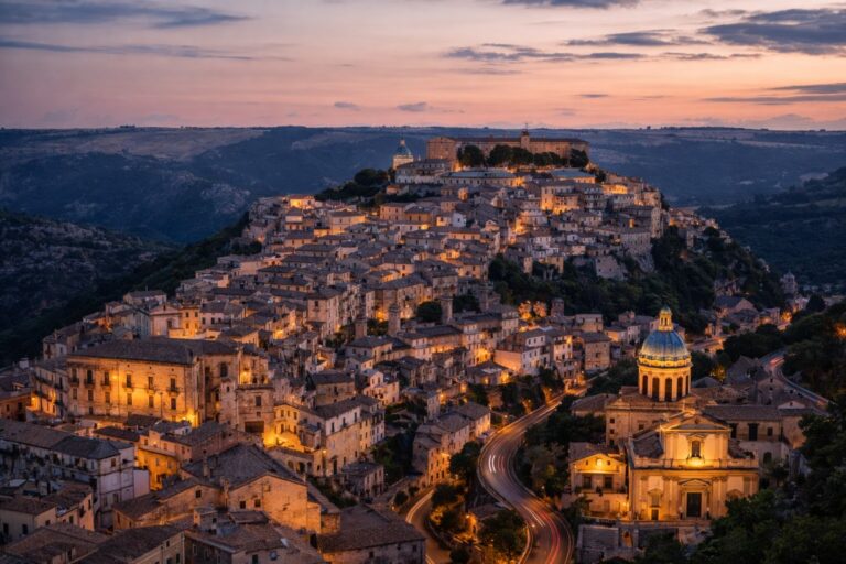 Ragusa Ibla at dusk with layered limestone buildings climbing a hill and warm streetlights tracing the winding road.